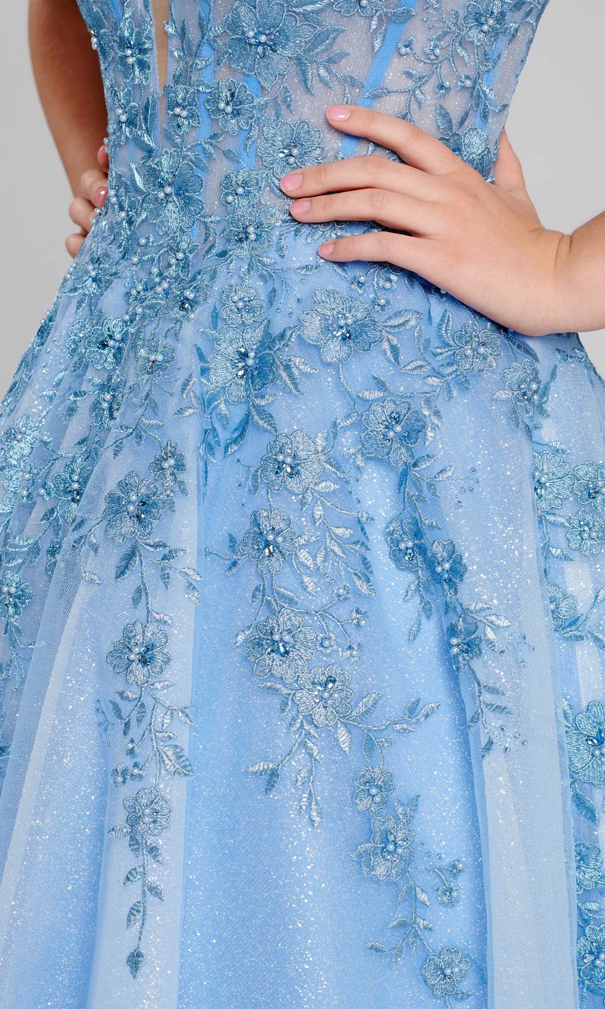 Close-up of a person wearing the Long Glitter Prom Ball Gown: Ellie Wilde EW120014 in light blue, featuring intricate floral embroidery, sequins, and glitter tulle. One hand rests on the waist.
